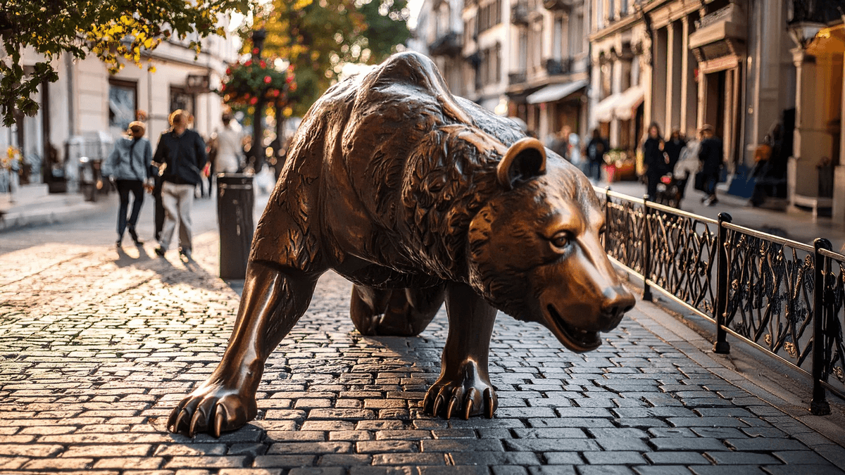 Large bronze bear statue crouched on a cobblestone street, with blurred historic buildings and pedestrians in the background under warm golden light.