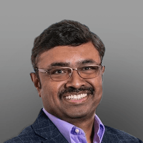 Professional headshot of a smiling man with dark hair, mustache, and glasses, wearing a patterned blazer and purple shirt.