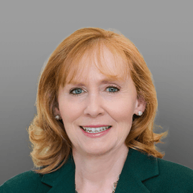 Smiling woman with reddish-blonde hair and blue eyes wearing a dark green blazer.