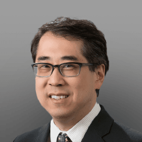 Smiling professional man of East Asian descent wearing black-rimmed glasses, a dark suit, and a patterned tie.