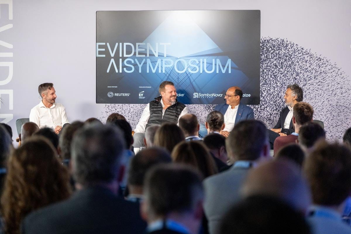 Four panelists discussing on stage at the Evident AI Symposium, with sponsor logos for Reuters, EY, Snowflake, and SambaNova Systems visible on the screen behind them.