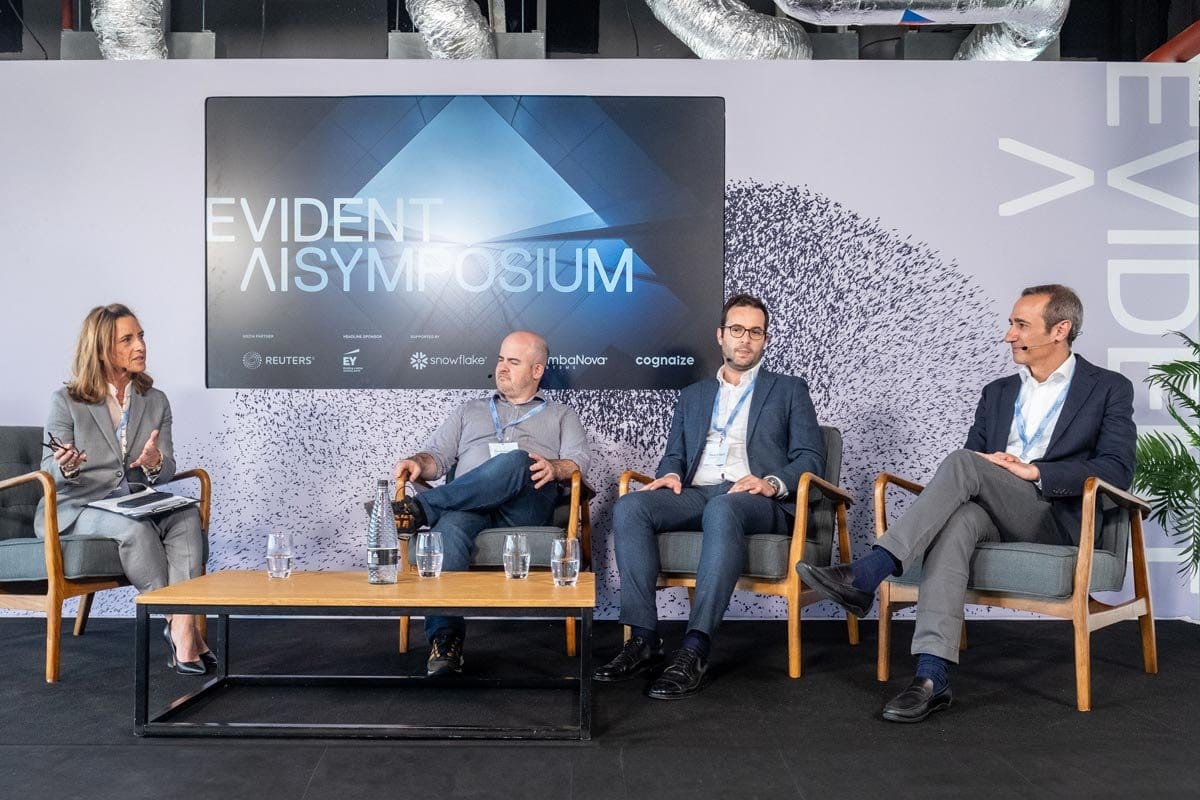 Panel discussion at the Evident AISYMPOSIUM, showing a woman speaking and three men listening, against a backdrop featuring the event title and sponsor logos like Reuters, EY, Snowflake, mbaNova, and Cognaize.