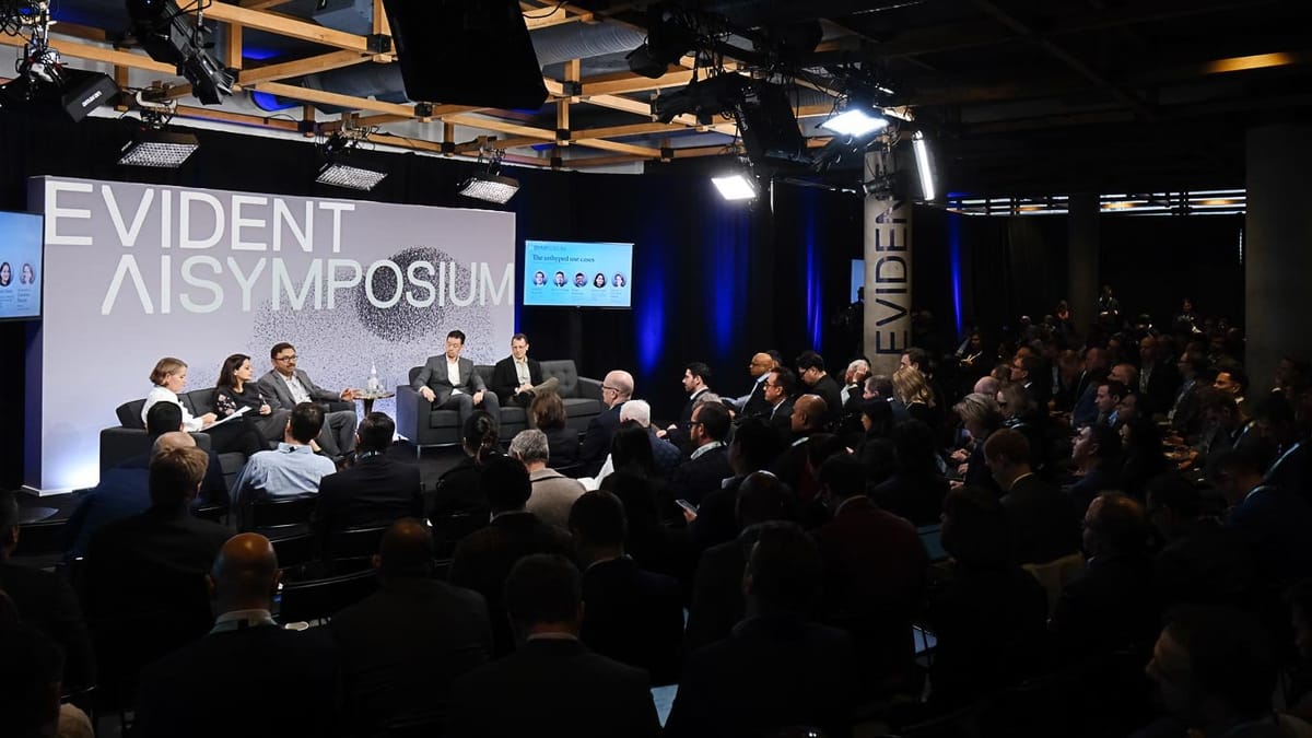 A diverse panel of six speakers sits on a stage, discussing at the Evident AI Symposium, with a large audience observing. A prominent backdrop banner reads 'EVIDENT AI SYMPOSIUM' and screens display relevant event content.
